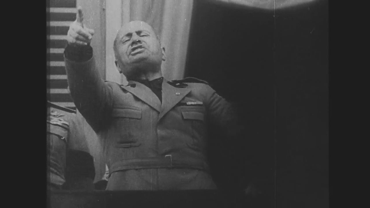 Un chef d'État des années 1930 harangue la foule, le bras tendu depuis un balcon