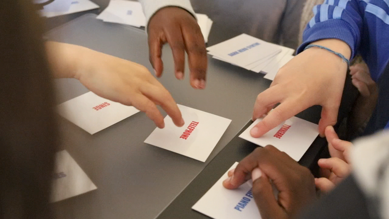 Des mains d'élèves attrapent les cartes-mots TÉLÉPHONE, SINGE, PIANO posées sur la table